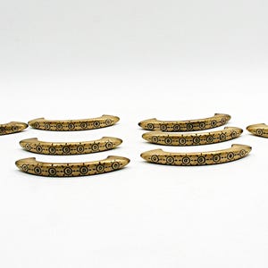 May include: A collection of ten curved, gold-toned metal drawer pulls. Nine of the pulls feature a repeating circular pattern with a central dot, while one pull displays a distinct wavy line design. Each piece has a smooth, metallic finish.