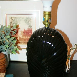 Può includere: Una lampada da tavolo decorativa verde scuro con una lampadina. La lampada ha una base strutturata con un design a spirale. La lampada è su una superficie scura, con un'immagine incorniciata e vegetazione sullo sfondo.