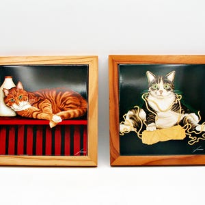 May include: Two framed cat art pieces. One features a ginger tabby cat resting on a red and black striped surface with a milk bottle. The other shows a black and white cat with yarn. Both are in wooden frames.