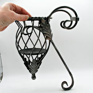 Peut inclure: Un bougeoir en métal foncé orné, avec un design décoratif en spirale. Le support présente une structure en forme de cage avec une base circulaire pour une bougie. La conception comprend des accents de feuilles et un bras incurvé pour suspendre.