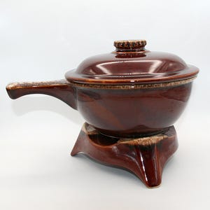 May include: A brown ceramic casserole dish with a lid and handle. The dish sits on a matching stand with three legs. The handle and stand have a lighter, speckled design.