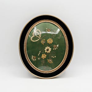 May include: An oval wall hanging featuring a green fabric background with embroidered gold and white flowers and a butterfly. The piece is encased in a black frame with a gold-toned, serrated inner border and a smooth outer gold-toned rim.