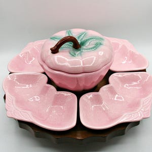May include: A pink ceramic serving set with a central lidded dish shaped like a tomato, surrounded by six matching serving dishes. The lid has a brown stem and green leaf details. The set is on a dark wooden tray.