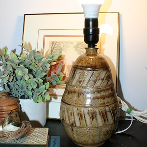 May include: A table lamp with a pear-shaped, speckled brown ceramic base featuring horizontal bands and diagonal etched lines. The lamp has a black socket and a bright white light bulb. The power cord is white and extends from the base.