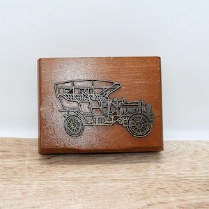 Può includere: Una scatola di legno rettangolare con un rilievo metallico dettagliato di un'auto d'epoca. L'auto è di colore metallico scuro ed è posta contro la calda venatura del legno marrone. La scatola è su una superficie di legno.