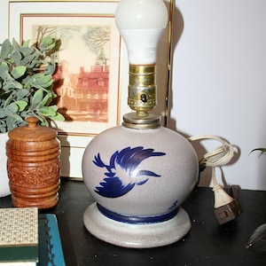 May include: A ceramic table lamp with a round, gray base and a blue bird design. The lamp has a white light bulb and a brass-colored socket. The lamp is on a black surface.