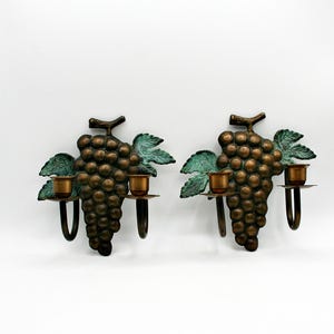 Może przedstawiać: Para ściennych kinkietów w kolorze brązu w kształcie kiści winogron, każdy z dwoma świecznikami. Winogrona są w kolorze ciemnego brązu, z zielonymi liśćmi z patyną. Świeczniki są w jaśniejszym kolorze brązu.