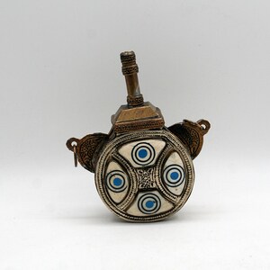 Könnte beinhalten: Ein antiker, verzierter Metallbehälter mit einer kreisförmigen, elfenbeinfarbenen Oberfläche. Die Oberfläche weist vier blau-weiße Kreise und ein zentrales Design auf. Ein messingfarbener Ausguss und dekorative Elemente vervollständigen das Design.
