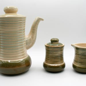 Peut inclure: Service à café en céramique beige et vert. L'ensemble comprend une cafetière avec couvercle, un sucrier avec couvercle et une crémière. Les pièces ont une texture côtelée et une base vert foncé.