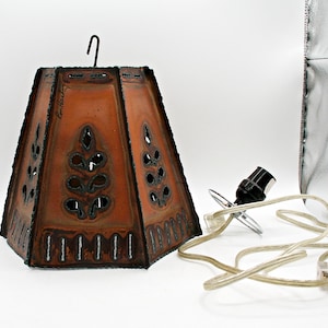 Peut inclure: Un abat-jour trapézoïdal en cuivre avec une patine sombre, des motifs de feuilles découpés et un crochet métallique noir sur le dessus. L'abat-jour est accompagné d'une douille noire et d'un cordon électrique transparent avec une prise.