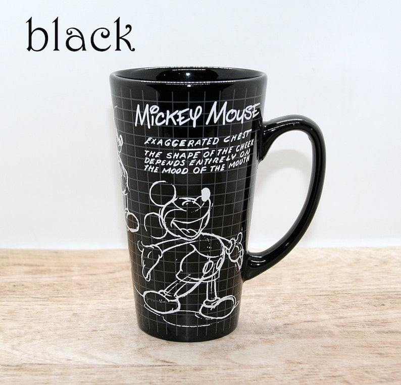 May include: Black ceramic Mickey Mouse mug with a large handle. The mug features a white outline drawing of Mickey Mouse and text that reads "MICKEY MOUSE" and "EXAGGERATED CHEST...THE MOOD OF THE MOUTH". The mug is tall and cylindrical.