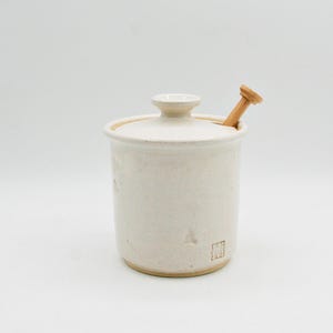 May include: A cream-coloured ceramic jar with a lid and a small wooden spoon. The jar has a cylindrical shape with a slightly textured surface. The lid has a small knob on top. The wooden spoon is inserted into the jar.