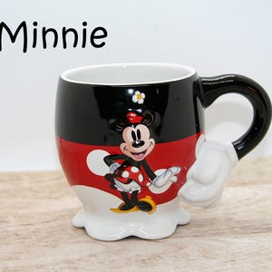 May include: A ceramic Minnie Mouse mug with a black handle and a white glove-shaped accent. The mug features a black top, red middle, and white bottom, with a Minnie Mouse graphic. The word "Minnie" is written in black at the top.