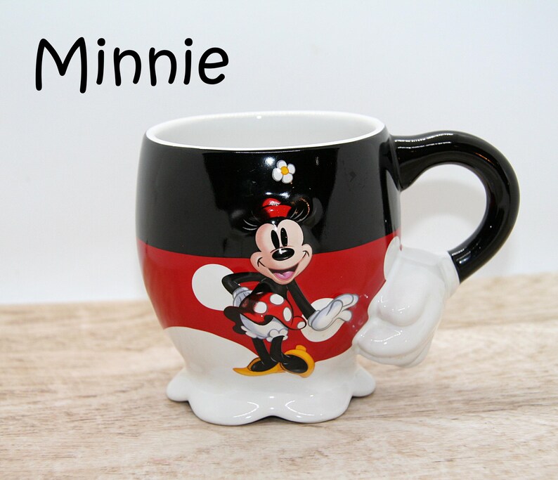 May include: A ceramic Minnie Mouse mug with a black handle and a white glove-shaped accent. The mug features a black top, red middle, and white bottom, with a Minnie Mouse graphic. The word "Minnie" is written in black at the top.