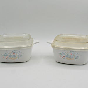 May include: Two white casserole dishes with clear lids. Each dish features a floral design with blue ribbons and red flowers. The dishes have handles on each side. The lids are slightly tinted.
