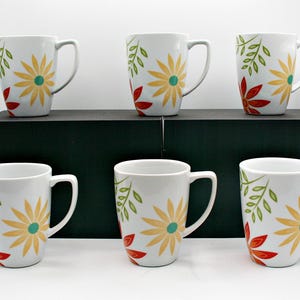 May include: A set of six white ceramic mugs, each featuring a repeating pattern of yellow flowers with teal centres, red flower petals, and green leafy sprigs. The mugs have a standard cylindrical shape with a curved handle on the right side.