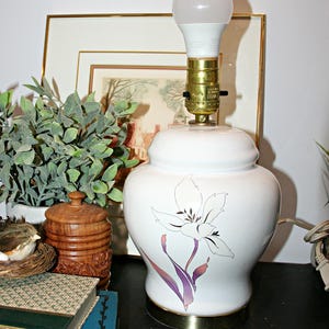 May include: White ceramic table lamp with a floral design in purple and black. The lamp has a brass base and a light bulb. The lamp is on a black surface, with a framed picture and greenery in the background.