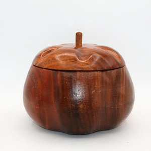 May include: A round, dark brown wooden container with a lid shaped like a pumpkin. The wood features a visible grain pattern and a smooth, polished finish. A small, cylindrical wooden stem is centered on top of the lid, which has wavy edges.