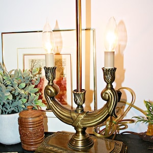 Op de afbeelding: Een koperkleurige metalen kandelaarlamp met een rechthoekige voet met versierde poten, een centrale verticale stang en twee gebogen armen die lampen met vlamvormige glazen kappen vasthouden. De lamp heeft een gepolijste afwerking.