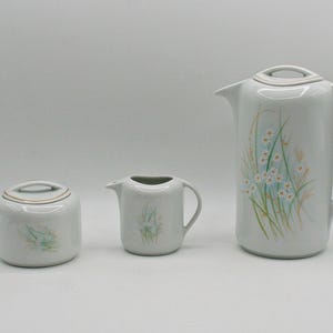 May include: A white ceramic tea set featuring a tall pitcher, a creamer, and a sugar bowl with a lid. Each piece is decorated with a floral design in shades of blue, green, and beige. The set is ideal for serving tea or coffee.