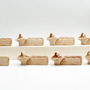 Könnte beinhalten: Ein Set aus acht beigen Keramik-Platzkartenhaltern, die jeweils ein kleines Korbmotiv auf einem rechteckigen Sockel mit geprägten Blumendetails an den Rändern aufweisen, angeordnet in zwei Reihen zu je vier auf einem weißen Block.