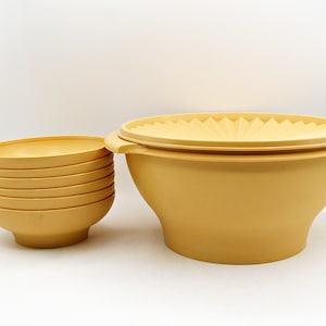 Può includere: Un set di ciotole vintage in plastica gialla. Viene mostrata una grande ciotola con coperchio e una pila di ciotole più piccole. Il coperchio ha un design decorativo a coste. Le ciotole sono realizzate in plastica resistente e leggera.