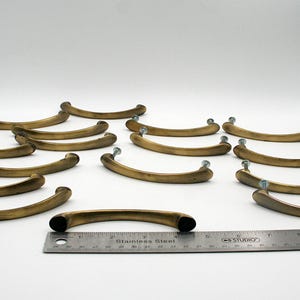 Op de afbeelding: Een verzameling antieke messingkleurige kastgrepen. Elke greep heeft een gebogen ontwerp met montageschroeven. Een roestvrijstalen liniaal is inbegrepen voor de schaal. Ideaal voor meubelrestauratie.