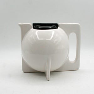 May include: A white ceramic teapot with a spherical body, a flat base, and a rectangular handle. The teapot features a small spout on the left side and a glossy black lid centred on top. The design is smooth with a clean, solid white finish.