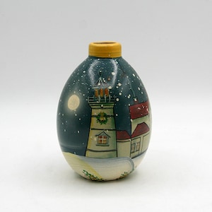 May include: A small, egg-shaped ceramic vase with a yellow rim, featuring a hand-painted scene of a lighthouse and adjacent buildings at night. The dark blue background includes a pale moon and white specks representing falling snow.