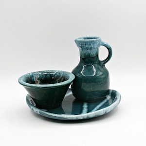 May include: A ceramic serving set in shades of teal and dark green. The set includes a small pitcher with a handle, a small bowl, and an oval-shaped tray. The glaze has a glossy finish with a mottled appearance.