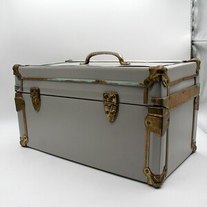 May include: A gray vintage trunk with brass hardware, including corner protectors, latches, and a handle. The trunk has a rectangular shape and is likely used for storage or decoration. The trunk's dimensions are approximately 24 inches long, 12 inches wide, and 12 inches high.