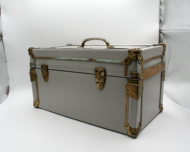 May include: A gray vintage trunk with brass hardware, including corner protectors, latches, and a handle. The trunk has a rectangular shape and is likely used for storage or decoration. The trunk's dimensions are approximately 24 inches long, 12 inches wide, and 12 inches high.