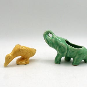 Pode incluir: Duas figuras de cerâmica: um pato amarelo com a cabeça baixa e um elefante verde com a tromba levantada. O elefante tem as costas escavadas, possivelmente para guardar pequenos objetos. Ambos estão sobre uma superfície branca.