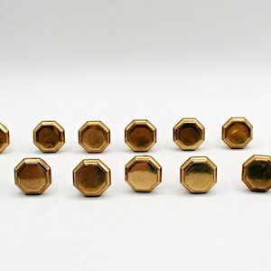 May include: A collection of thirteen brass-toned metal cabinet knobs, each featuring a distinct octagonal shape with a smooth, flat top surface and beveled edges, arranged in two rows against a plain, neutral background.