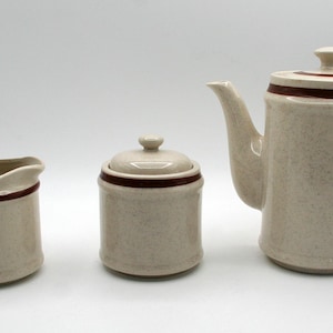 May include: A three-piece ceramic coffee set in a speckled beige finish with a dark brown trim. The set includes a creamer, a lidded sugar bowl, and a coffee pot with a handle and spout. The coffee pot is approximately 9 inches tall.