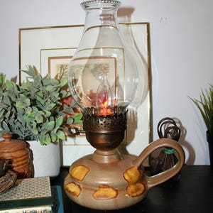 May include: A vintage oil lamp with a clear glass globe and a brown ceramic base. The base has a handle and is decorated with yellow-brown spots. The lamp is lit, with a warm, orange glow. The lamp is on a black surface.
