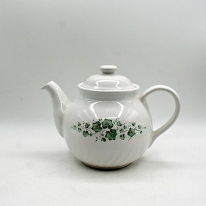May include: White ceramic teapot with a lid and handle. The teapot has a green ivy leaf pattern around the body. The spout and handle are curved. The teapot is designed for brewing tea.