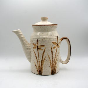 May include: Cream ceramic teapot with a lid and handle. The teapot has a brown dragonfly and grass design. The spout and handle are brown. The teapot has a speckled finish and a ribbed texture.