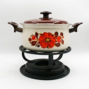 May include: A cream-colored enamel pot with a speckled finish, featuring a large red floral design on the front. The pot has a brown lid, two brown side handles, and sits on a black metal stand with a circular base and curved supports.