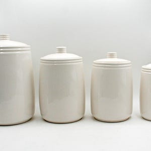 May include: A set of four cylindrical off-white ceramic canisters of varying heights, each featuring a smooth glossy finish, a rounded base, and a matching lid with a small knob handle at the top, arranged in a row from largest to smallest.
