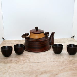 Peut inclure: Une théière en bois brun foncé avec une bande de bois clair autour du centre, un couvercle assorti et un bec verseur incurvé. La théière est accompagnée de quatre petites tasses en bois brun foncé assorties disposées devant elle.