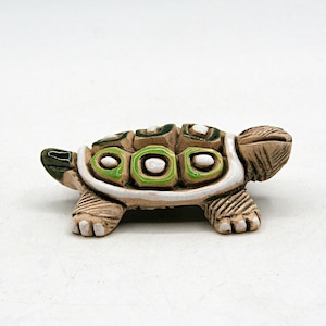 May include: A small ceramic turtle figurine with a textured tan body and a shell featuring raised, glossy green and white geometric accents. The turtle has a defined head, four legs with visible claw details, and a small tail, all finished in a matte tan glaze.