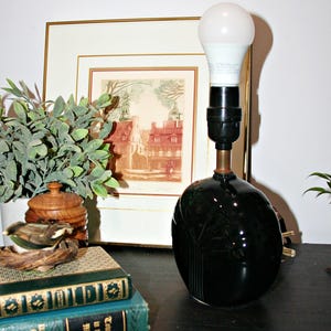 May include: A table lamp with a glossy, dark green, rounded ceramic base featuring a subtle embossed tree design. The lamp has a black socket and a white LED bulb. It stands next to two stacked green books and a small wooden lidded container.