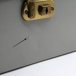 May include: Close-up of a gray metal box with a brass-colored latch. The box has a small dent and a black arrow pointing to it. The latch is rectangular with a keyhole.