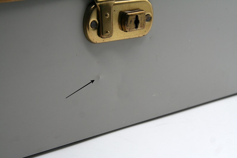 May include: Close-up of a gray metal box with a brass-colored latch. The box has a small dent and a black arrow pointing to it. The latch is rectangular with a keyhole.