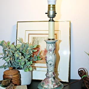 May include: A table lamp with a cream-colored ceramic base featuring a floral pattern in pink, green, and gold. The lamp has a brass-colored metal socket and a white light bulb. It sits on a dark brown circular wooden base.