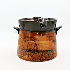 May include: A ceramic pot with a lid and two side handles. The upper portion is finished in a glossy dark brown glaze, while the lower body features a textured, mottled orange and brown glaze with visible dark brown streaks and speckles.