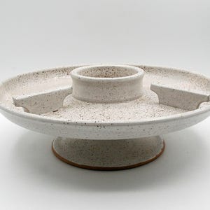 May include: A round ceramic serving platter with a speckled off-white glaze. The platter features a central raised circular bowl and two small rectangular dividers on the surface, all supported by a wide, sturdy pedestal base with a brown rim.