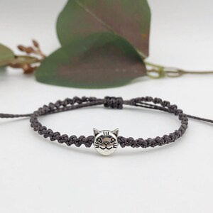May include: A dark grey macramé bracelet with a silver cat charm. The bracelet is adjustable with sliding knots and is displayed on a white surface. The cat charm has etched details.