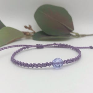 May include: A lavender macrame bracelet with a central, light purple, faceted bead. The bracelet is adjustable. The macrame knotting is visible. A delicate piece of jewellery. The bracelet is a soft lavender colour.